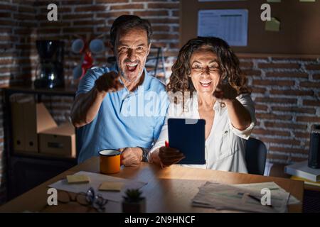 Middle age hispanic couple using touchpad sitting on the table at night pointing to you and the camera with fingers, smiling positive and cheerful Stock Photo