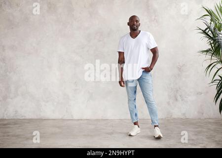 Handsome African American young man stands full body, looks at camera, against bright textured wall in studio. Male model wears casual clothes white T Stock Photo