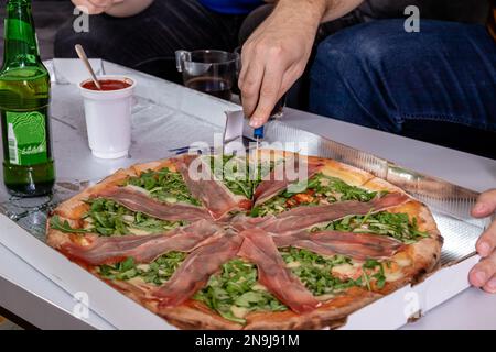Fresh pizza with slices of ham and arugula leaves. A hand holding a knife and cutting slices from a delivery box. Home party. Stock Photo