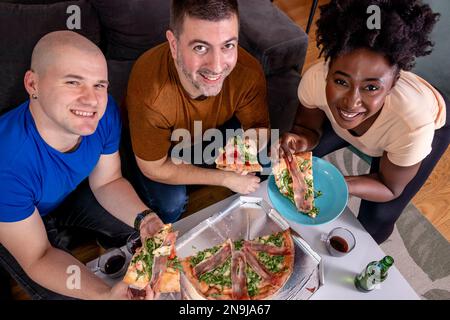 Group of young friends eating pizza. Home party. High angle view. Looking at the camera. Stock Photo