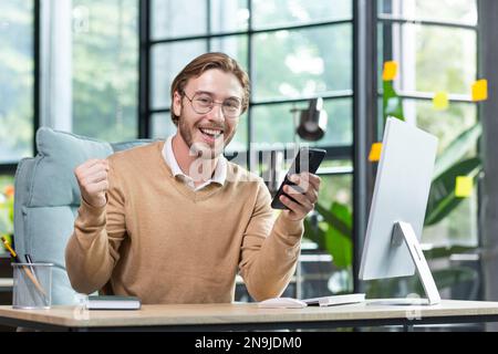 Happy young male programmer, freelancer sits in the office, holds the phone, shows a victory gesture with his hand yes. Received good news, messages, playing online games, successful transaction. Stock Photo
