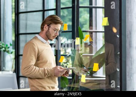 Portrait of a young programmer standing in the office near a glass transparent board and working with concentration on a tablet. Planning, developing, typing. Stock Photo