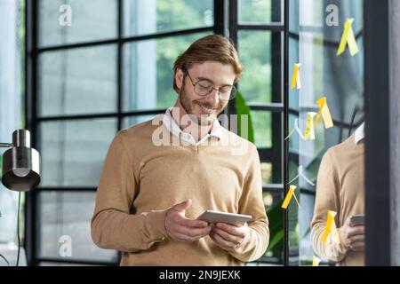 Portrait of a young smiling and successful programmer standing in the office near a glass transparent board and working on a tablet. Plans, develops, typing, notes with notes on the board. Stock Photo