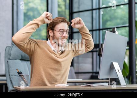 Smiling young male programmer, freelancer sits at the table, looks happy at the computer monitor. Raised his hands up, rejoices in success, victory. Stock Photo