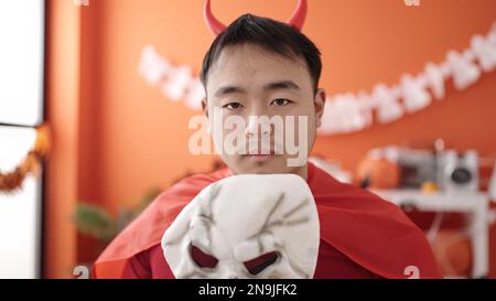 Young chinese man wearing devil costume holding skull mask at home Stock Photo
