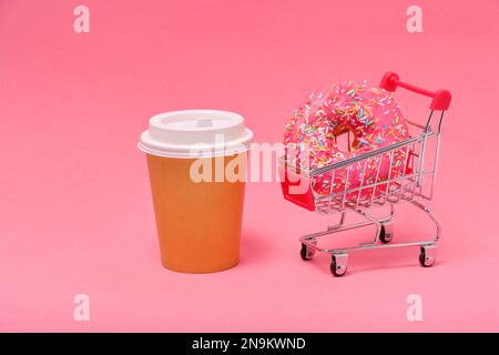 Pink donut in shopping cart and disposable coffee cup on pink background Stock Photo