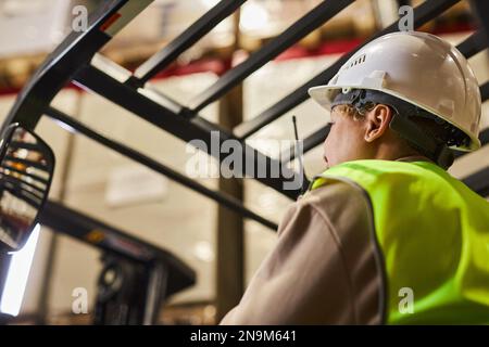 Female worker wearing hardhat while operating forklift truck in warehouse and talking to portable radio Stock Photo