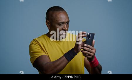 Athletic mature african american man in studio on blue background looking at mobile phone screen pensive male athlete sportsman in sportswear browsing Stock Photo