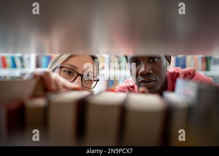 Closeup through bookshelf couple of students looking for right book in library to prepare for exam. Stock Photo
