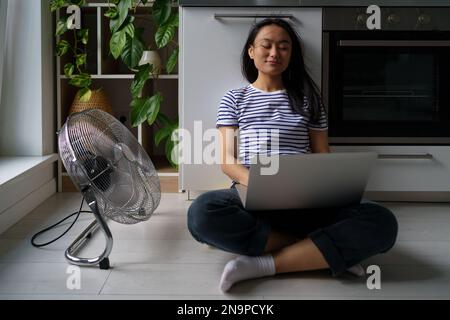 Contented Asian woman turn on floor ventilator fan while work with laptop joyful self save from heat Stock Photo