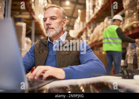 Bearded mature man using laptop in storage warehouse while managing distribution business Stock Photo