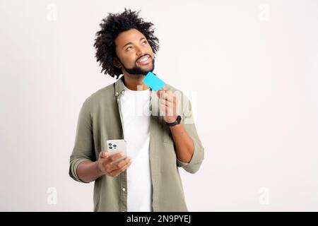 Inspired cheerful young african american man 30s holding smartphone and credit bank card isolated on white background, looking away lost in dreams, choosing something, making purchase online, shopping Stock Photo