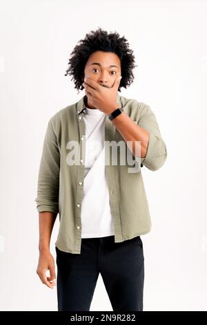 Amazed African-American young man is covering up his mouth and staring at the camera with wide opened eyes isolated on white background, shocked curly guy speechless Stock Photo