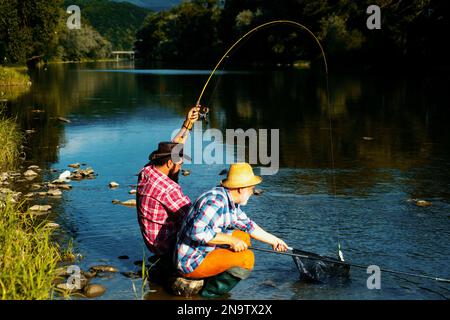 Male hobby fishing. Two male friends dressed in shirts fishing together with net and rod during the morning light on the lake. Man at riverside enjoy Stock Photo