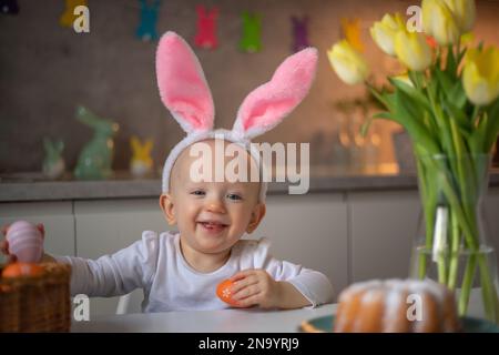 happy cute little baby girl wearing bunny ears on easter day sitting at the table in the kitchen. Stock Photo