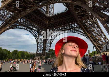 Young woman wearing a red hat stands under the Eiffel Tower and basks in the sunlight in Paris, France; Paris, France Stock Photo