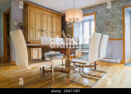 Antique oak wood table with white high back leather and chrome chairs in dining room with old wooden buffet and ash floorboards inside old 1820 home. Stock Photo