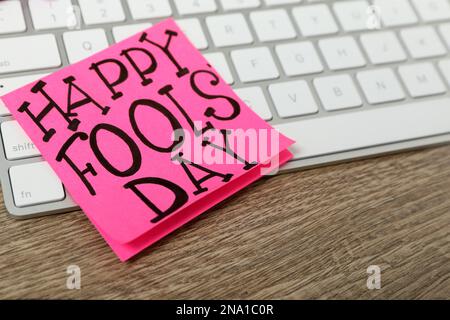 Note with phrase Happy Fools' Day and keyboard on wooden table, closeup Stock Photo