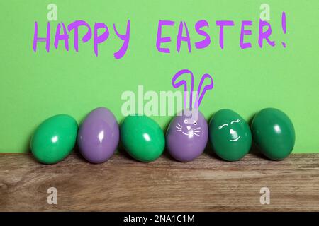 One egg with drawn face and ears as Easter bunny among others on wooden table against green background Stock Photo