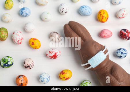Chocolate bunny with protective mask and eggs on white wooden table, flat lay. Easter holiday during COVID-19 quarantine Stock Photo
