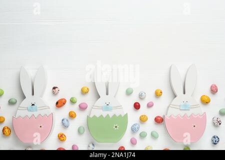Candies and bunny figures in protective masks on white wooden table, flat lay with space for text. Easter holiday during COVID-19 quarantine Stock Photo