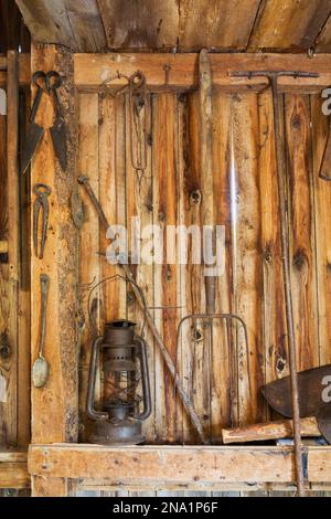 Antique oil lantern, pitchfork, shears, axe and other manual tools hanging on vertical wood plank wall inside old barn. Stock Photo