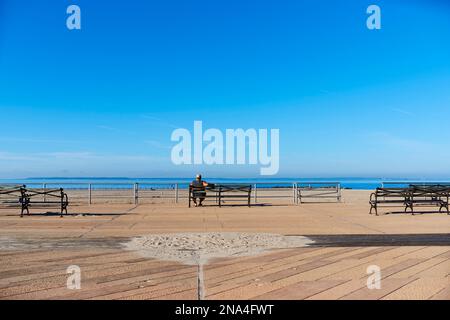 Man sitting on bench along Coney Island boardwalk looking out to the Atlantic Ocean; New York City, New York, United States of America Stock Photo