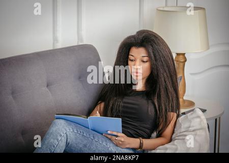A beautiful African-American girl with a notebook in her hands sits on the sofa and makes notes in a notebook. Stock Photo