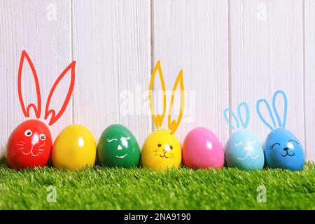 Several eggs with drawn faces and ears as Easter bunnies among others on green grass against white wooden background Stock Photo