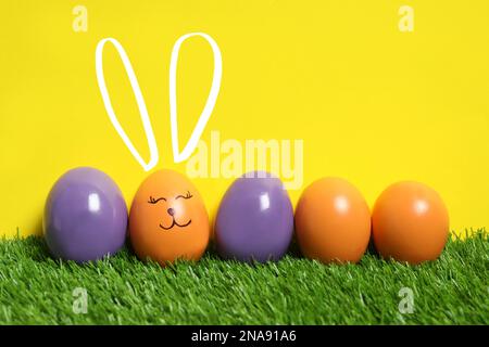 One egg with drawn face and ears as Easter bunny among others on green grass against yellow background Stock Photo