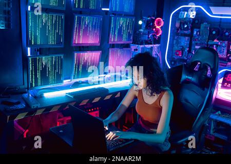 Female Asian hacker in hideout place with dark environment and multiple displays and cables, trying to break into data servers and infect the system with a virus.  Stock Photo