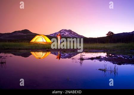 Man with a camera and tripod photographing Oregon beauty at sunrise next to tent at Sparks Lake, with South Sister and Broken Top in the background... Stock Photo