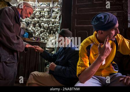 In the Old Medina of Fes, Morocco, there are hundreds of shops selling local wares. Behind one contemplative man, two gentlemen discuss price of pr... Stock Photo