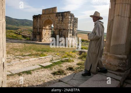 A Moroccan man dressed in traditional clothing walks on the grounds of Volubilis, Morocco; Volubilis, Morocco Stock Photo