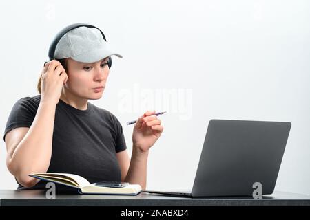 Busy young woman in headphones working on laptop, making video call and making notes in notebook, work and study online. Stock Photo