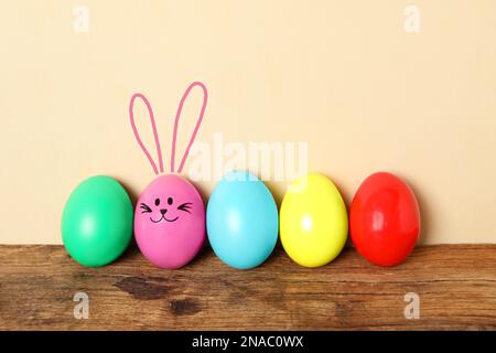 One eggs with drawn face and ears as Easter bunny among others on wooden table against beige background Stock Photo