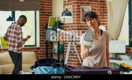 Smiling person singing and dancing while she irons clothing, housewife having fun doing chores in at home. Cheerful wife enjoying ironing clothes and listening to music. Handheld shot. Stock Photo