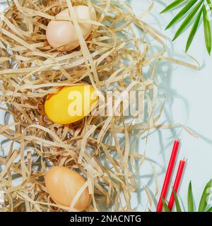 Easter holiday composition with colored eggs on shredded paper, red church candles and palm leaf on blue background. Springtime. Congratulatory easter Stock Photo