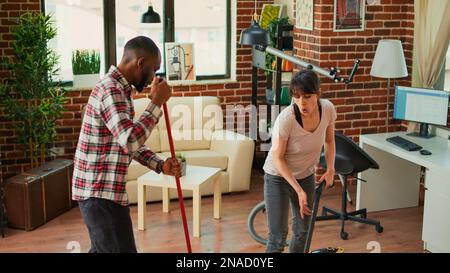 Smiling young people using tools to clean household, listening to music and dancing while they use mop and vacuum. Diverse couple cleaning apartment floors, sweeping dust with solution. Stock Photo