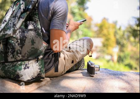 Cropped image of an Asian male traveler with backpack and film camera sits on a rock using his smartphone, resting while trekking. Stock Photo