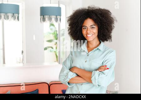 Ethnic woman looking at the camera and smiling standing in modern apartment, feeling comfort and relaxation. Cheerful African-American 20s young woman with arms crossed Stock Photo