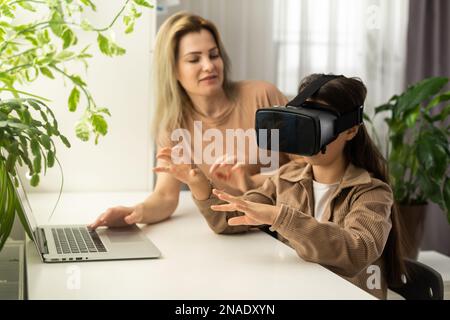 Blond excited mother helping her adorable girl teenager virtual using virtual reality headset at home interior. Stock Photo