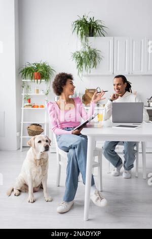 Smiling african american woman with notebook talking to boyfriend near devices and labrador at home Stock Photo