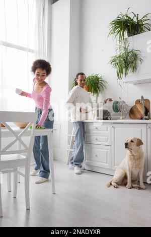 Positive african american woman holding cup and cleaning table near boyfriend and labrador in kitchen Stock Photo