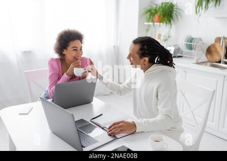 Cheerful african american couple talking near devices and coffee in kitchen Stock Photo