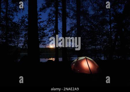 Night campsite with a illuminated tent and setting sun in forest. Stock Photo