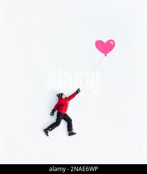 Aerial of boy laying on snow in winter clothing holding pink balloon. Stock Photo
