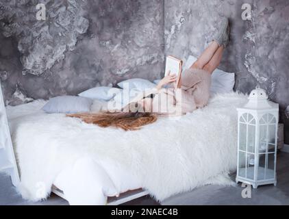 A long-haired blonde girl lies in bed and reads a book. Rest at home. Day off. Hobby and relaxation. Cozy interior. Study and reading Stock Photo
