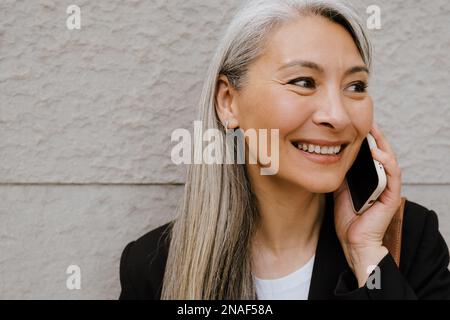 Grey asian woman smiling and talking on cellphone while standing by wall outdoors Stock Photo