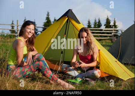 Two women hikers camping outdoors. Attractive, positive women hiking in summer. Female friends sitting on grass near tent, having break, smiling. Concept of tourism and adventure. Stock Photo